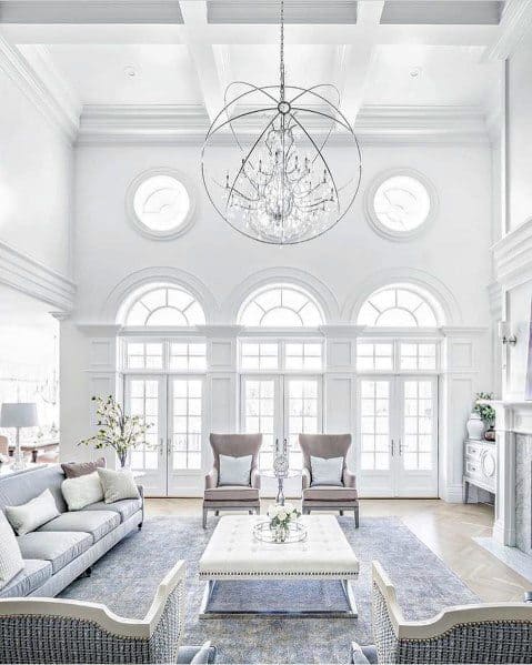 A bright, spacious living room featuring a coffered ceiling, elegant crown molding, large windows, and a luxurious chandelier