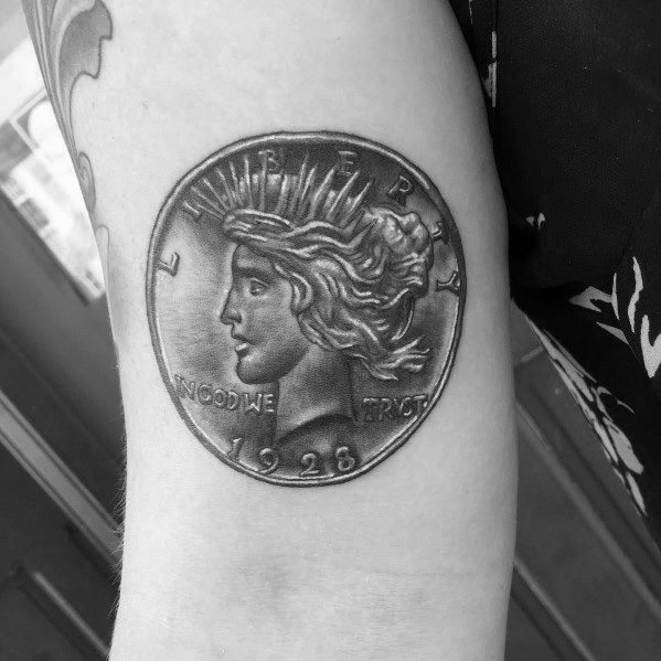 Detailed tattoo of a 1926 Peace Dollar coin on the arm, featuring Lady Liberty with intricate shading for a realistic, three-dimensional look.