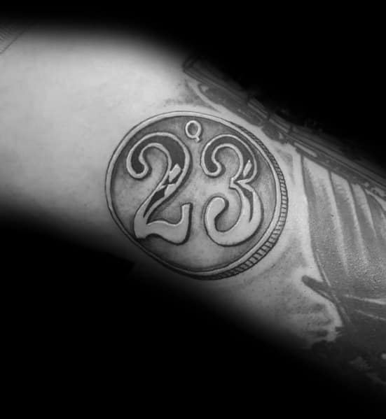 Minimalist tattoo of a coin with the number '23' in bold script, featuring fine shading and a classic rim design on the arm.