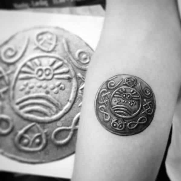 Intricate tattoo of an ancient-style coin with symbolic engravings, designed with realistic shading for a three-dimensional effect on the arm.