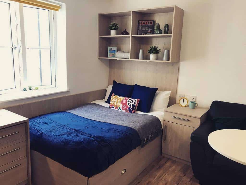 Simple college dorm bedroom with wooden furniture, navy bedding, and wall shelves for decor.