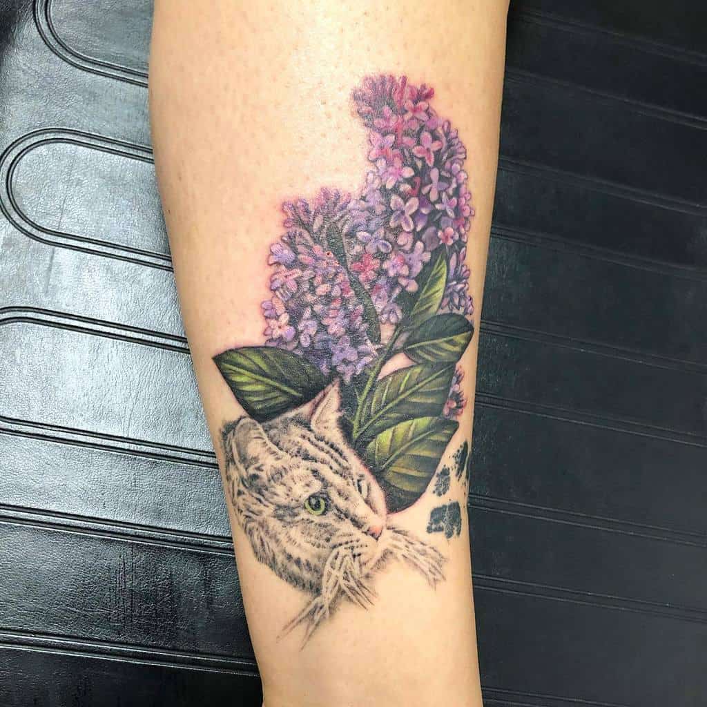 Realistic tattoo of a cat’s face surrounded by detailed lilac flowers and lush green leaves, blending fine shading and vibrant colors.