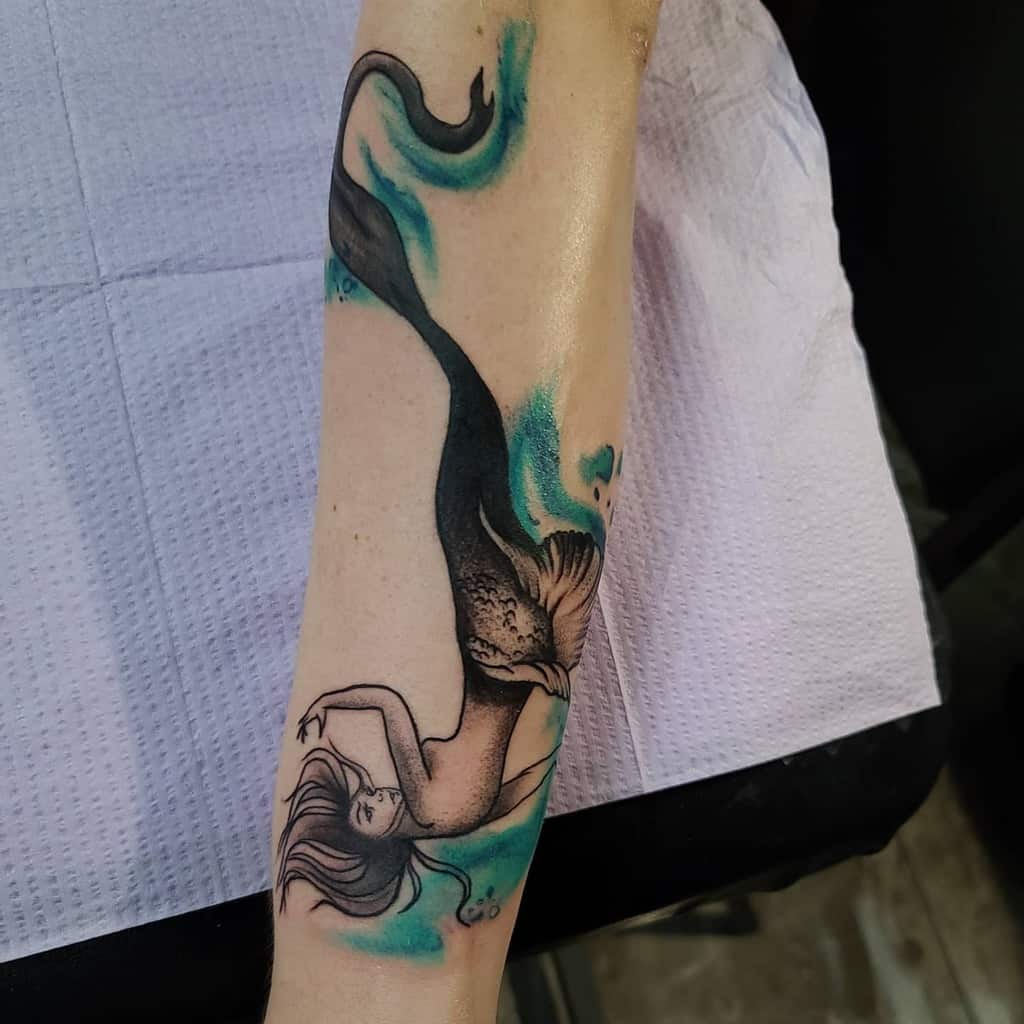 Tattoo of an upside-down mermaid with a dark, textured tail and blue watercolor accents, creating a fluid, surreal underwater effect.
