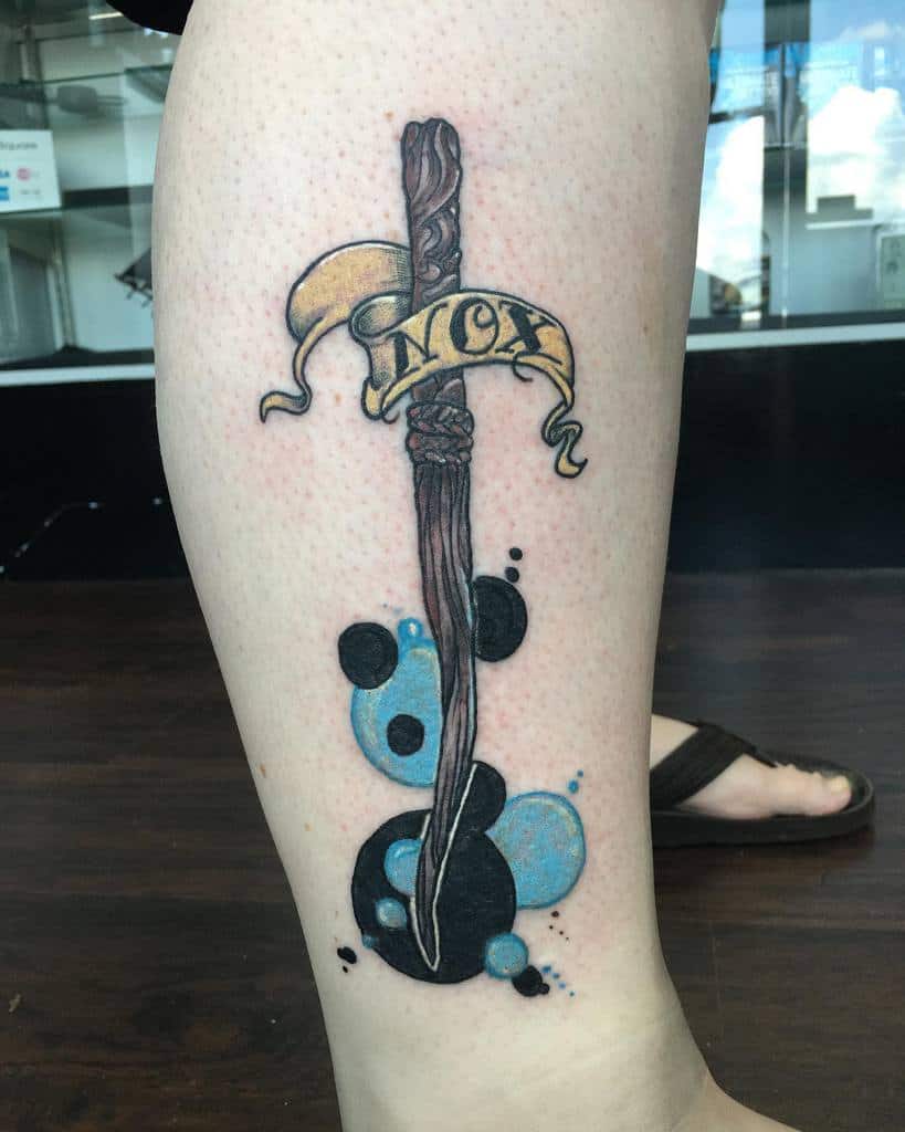 Harry Potter-inspired tattoo of a wand with a 'Nox' banner, featuring abstract blue and black ink splashes in a watercolor style