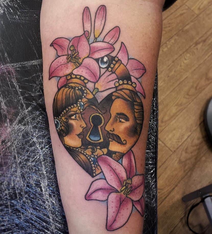 Traditional-style tattoo of a heart-shaped lock with a man and woman facing each other, surrounded by pink lilies