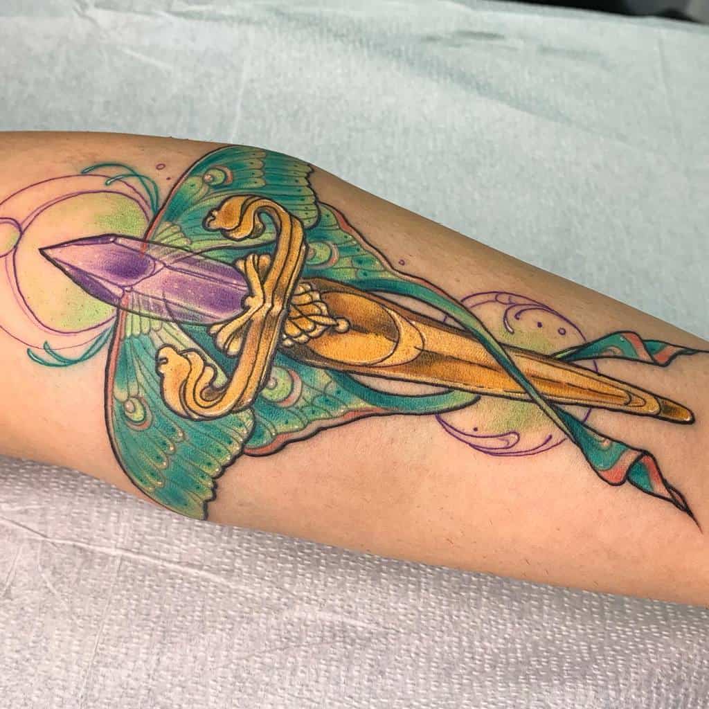 A colorful tattoo of a Luna Moth with a dagger piercing a purple crystal, surrounded by soft pastel swirls