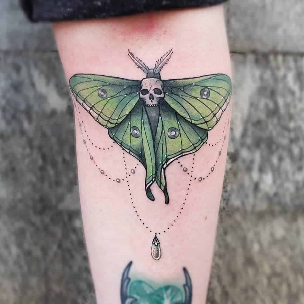 A colorful tattoo of a Luna Moth with green wings, a skull in the center, and hanging pearl-like chains with a drop pendant