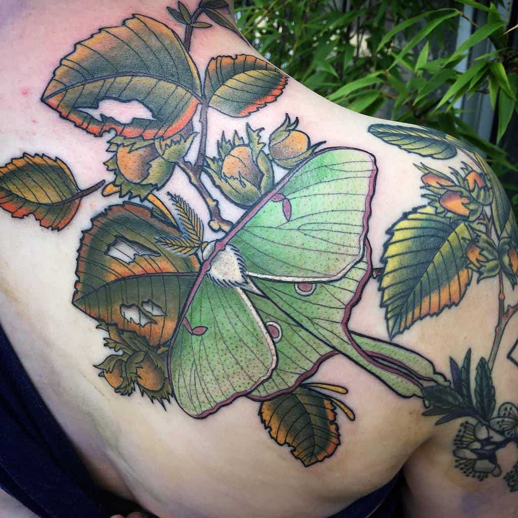 A vibrant tattoo of a Luna Moth, surrounded by detailed leaves and berries, with warm green, orange, and yellow tones
