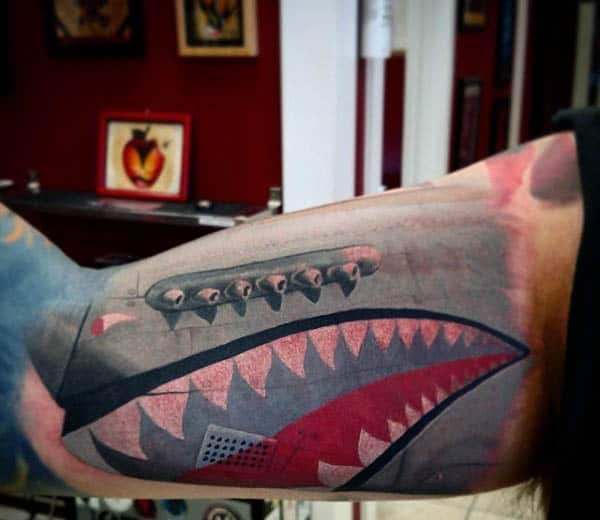 Tattoo of a shark face with teeth and gills on a person's upper arm, done in a colorful style, featuring an airplane soaring overhead