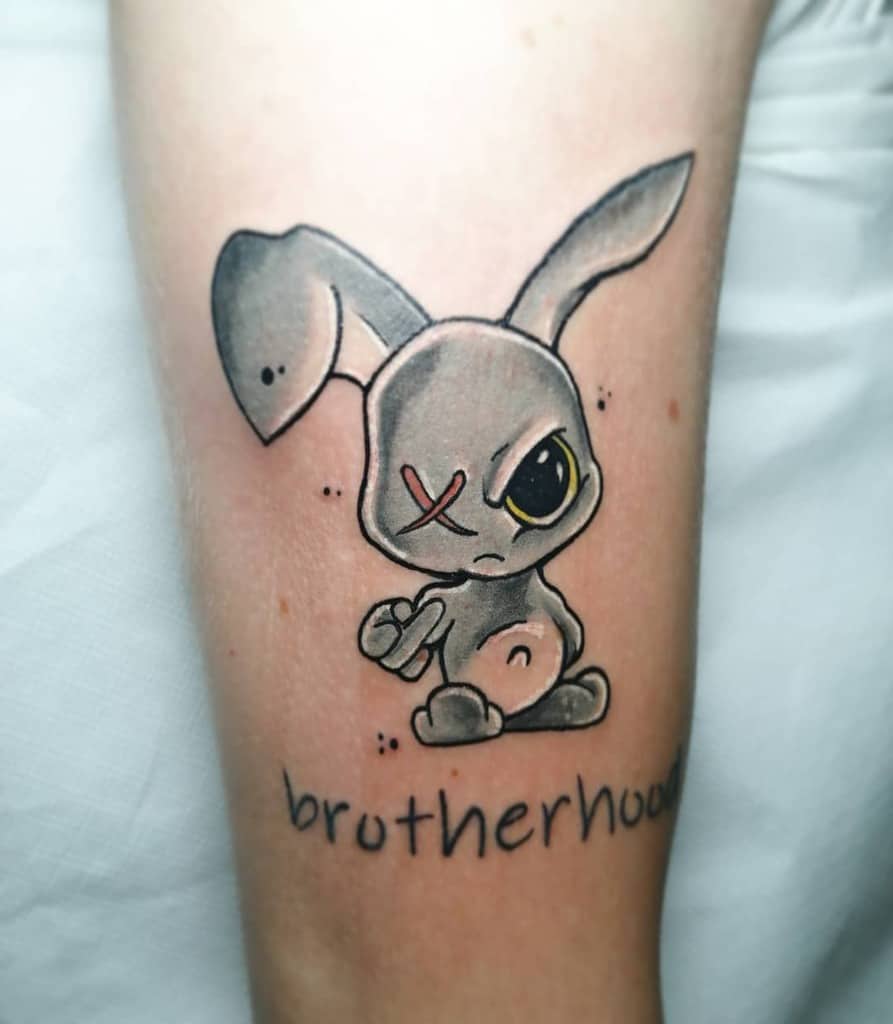 Playful tattoo of a gray rabbit with a stitched eye and yellow accent, holding a fist, with the word 'brotherhood' written underneath