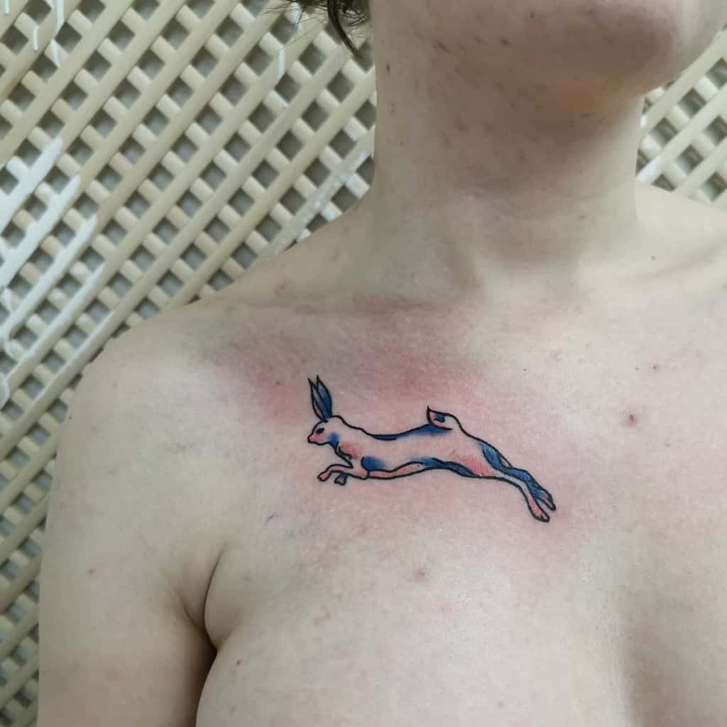 Minimalistic tattoo of a rabbit mid-leap, outlined in black with soft blue and pink accents, creating a fluid, dynamic movement