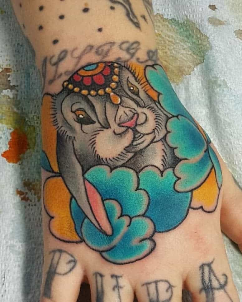 Colorful tattoo of a rabbit adorned with a jeweled headdress, surrounded by bold blue and orange clouds