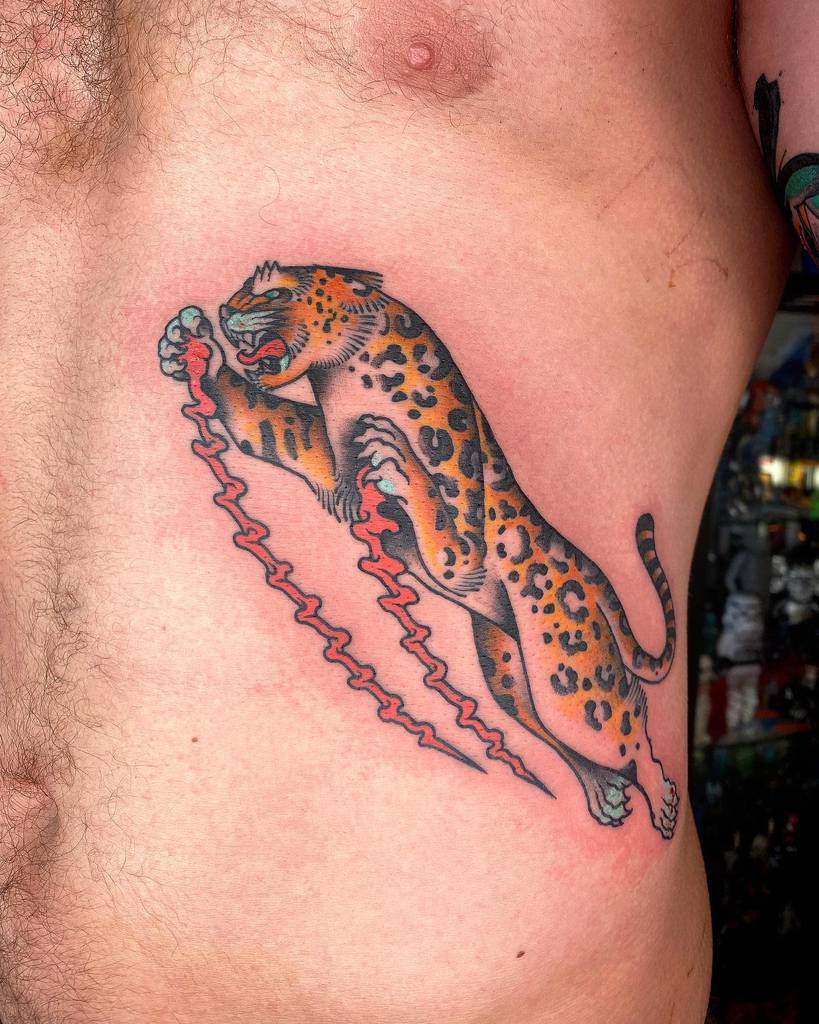 A bold traditional-style tattoo of a jaguar clawing down the ribs, featuring vibrant orange tones, fierce expression, and dynamic motion lines.
