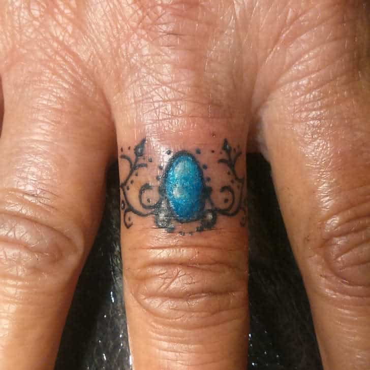 A delicate ring tattoo featuring a blue gemstone surrounded by ornate black swirls and dotted accents