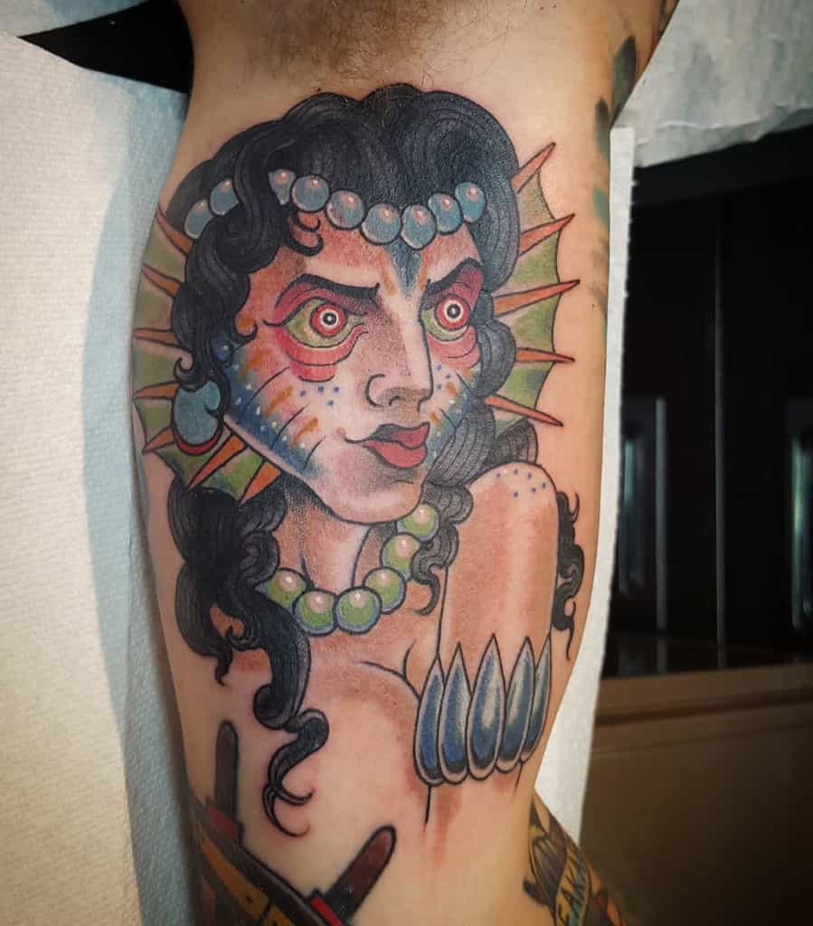 Vivid tattoo of a siren with striking red eyes, colorful scales, and detailed facial features, adorned with pearls and fins