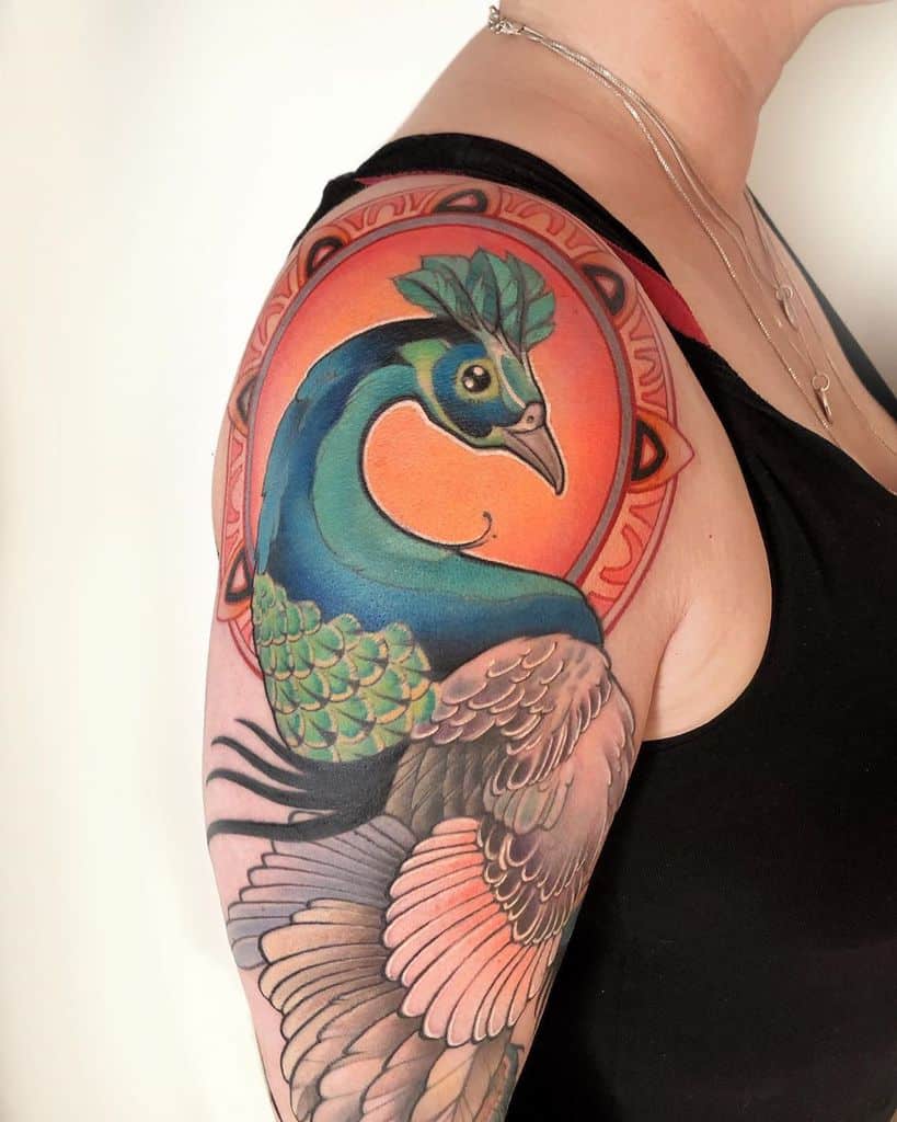 Colorful peacock tattoo with vibrant feathers and a sunburst background, wrapping around the shoulder.