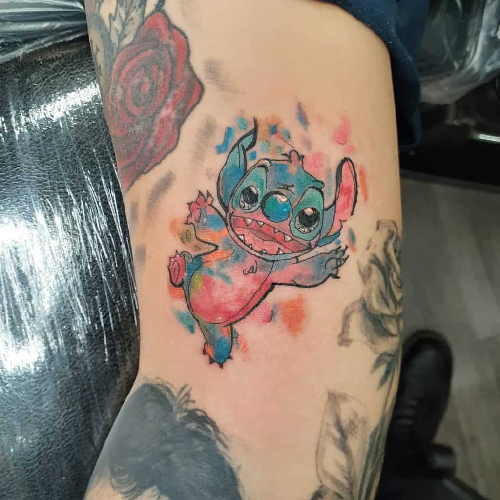 Colorful tattoo of Stitch from Lilo & Stitch with vibrant watercolor splashes, on the upper arm.