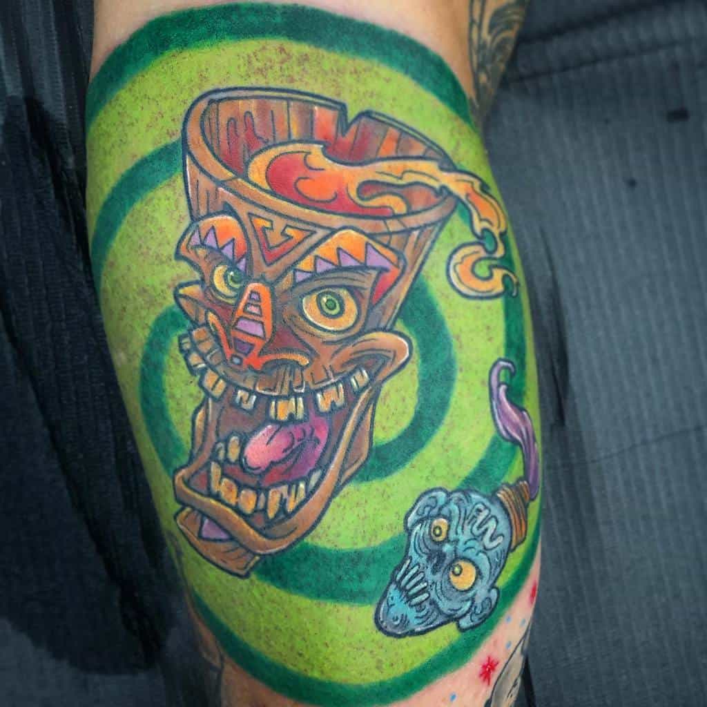 Colorful Tiki tattoo featuring a tiki mug with a grinning face and flames, surrounded by swirling green and yellow patterns, with a blue skull on the side