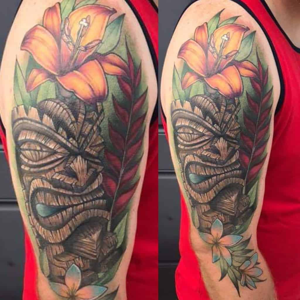 Vibrant Tiki tattoo, featuring a wooden Tiki mask surrounded by colorful flowers, leaves, with a mix of orange, green, and white tones