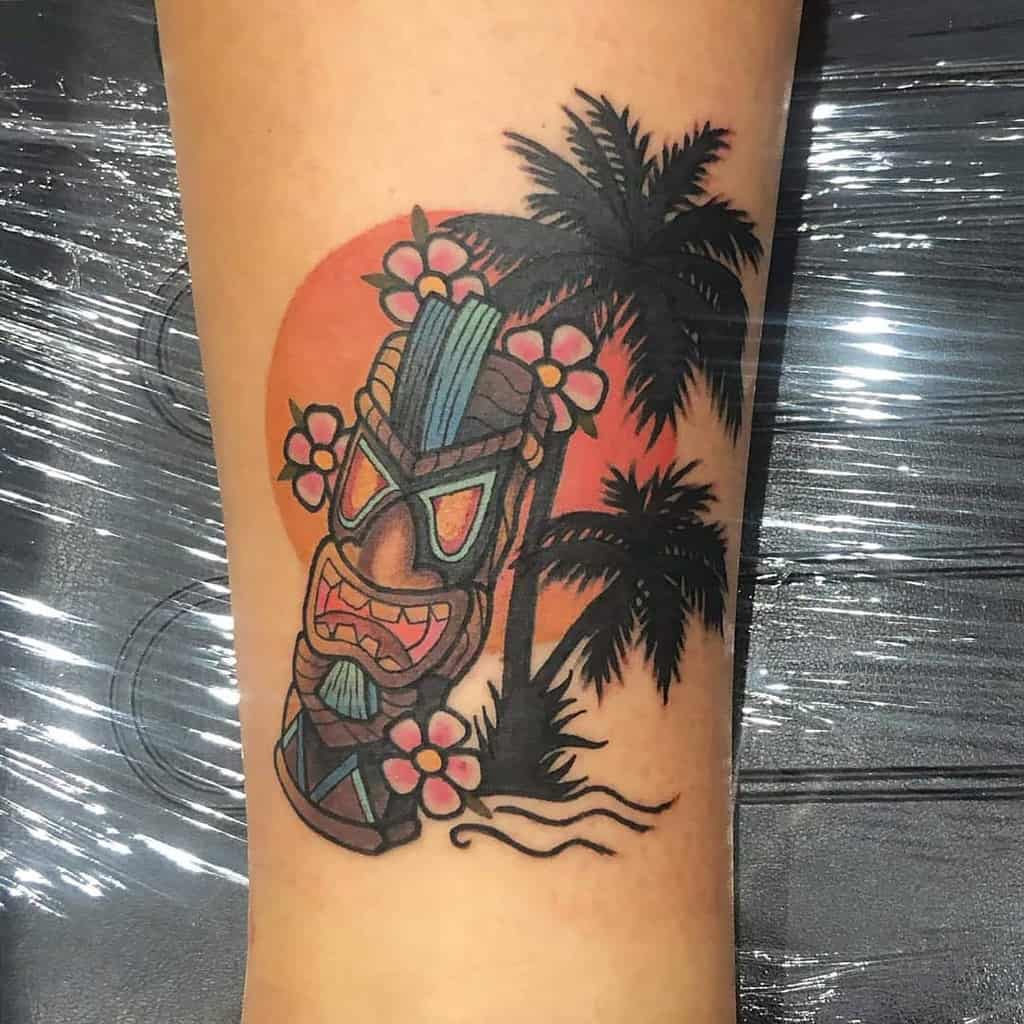 Colorful Tiki tattoo featuring a Tiki mask surrounded by tropical flowers, palm trees, and a sunset background, with vibrant orange, pink, and green tones