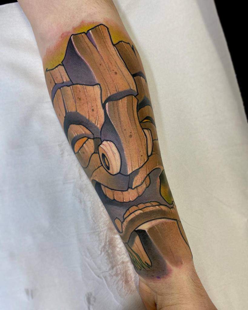 Colorful Tiki tattoo featuring a stylized wooden mask, bright highlights of yellow and green surrounding the face, and bold outlines