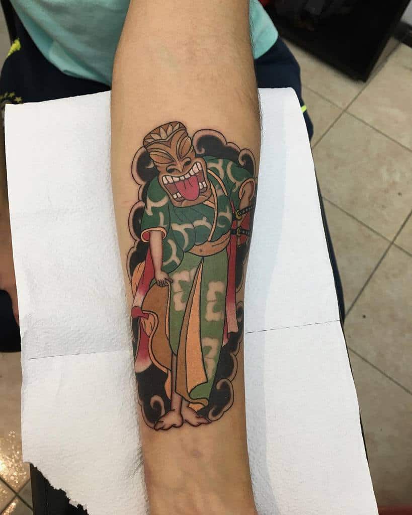 Colorful Tiki tattoo featuring a cartoonish Tiki mask character wearing traditional attire, with vibrant green, red, and yellow hues and black clouds