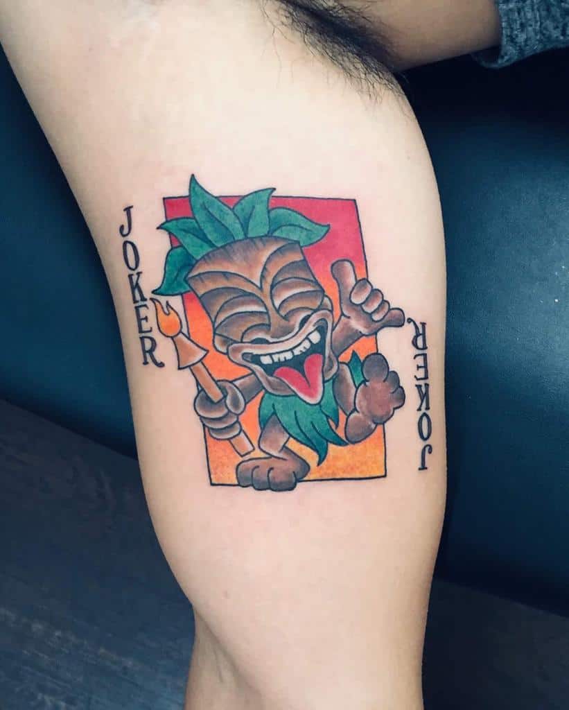 Colorful Tiki tattoo featuring a playful Tiki mask with a green leafy top, holding a staff, framed by a red-orange background and the word 'JOKER' in bold lettering