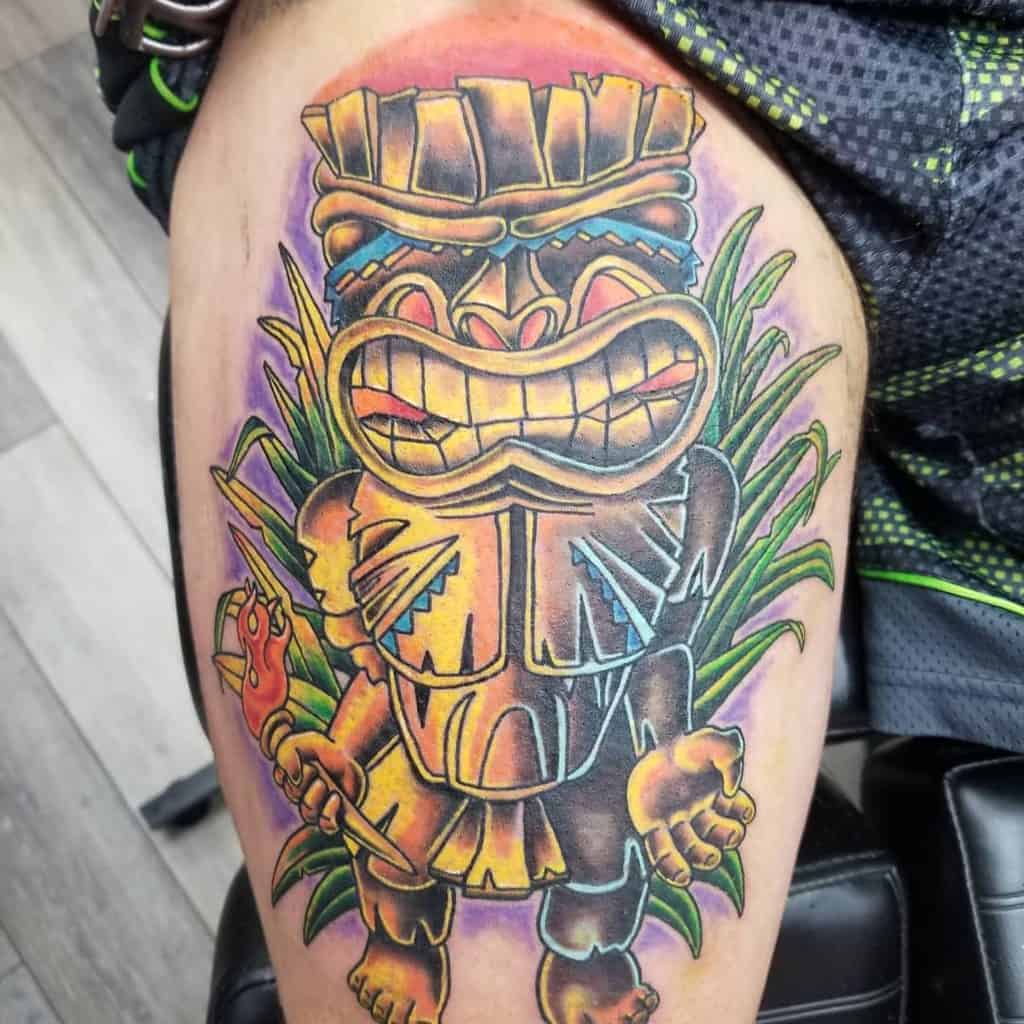 Vibrant Tiki tattoo featuring a bold, grinning Tiki mask with a colorful background of green plants and a torch in its hand