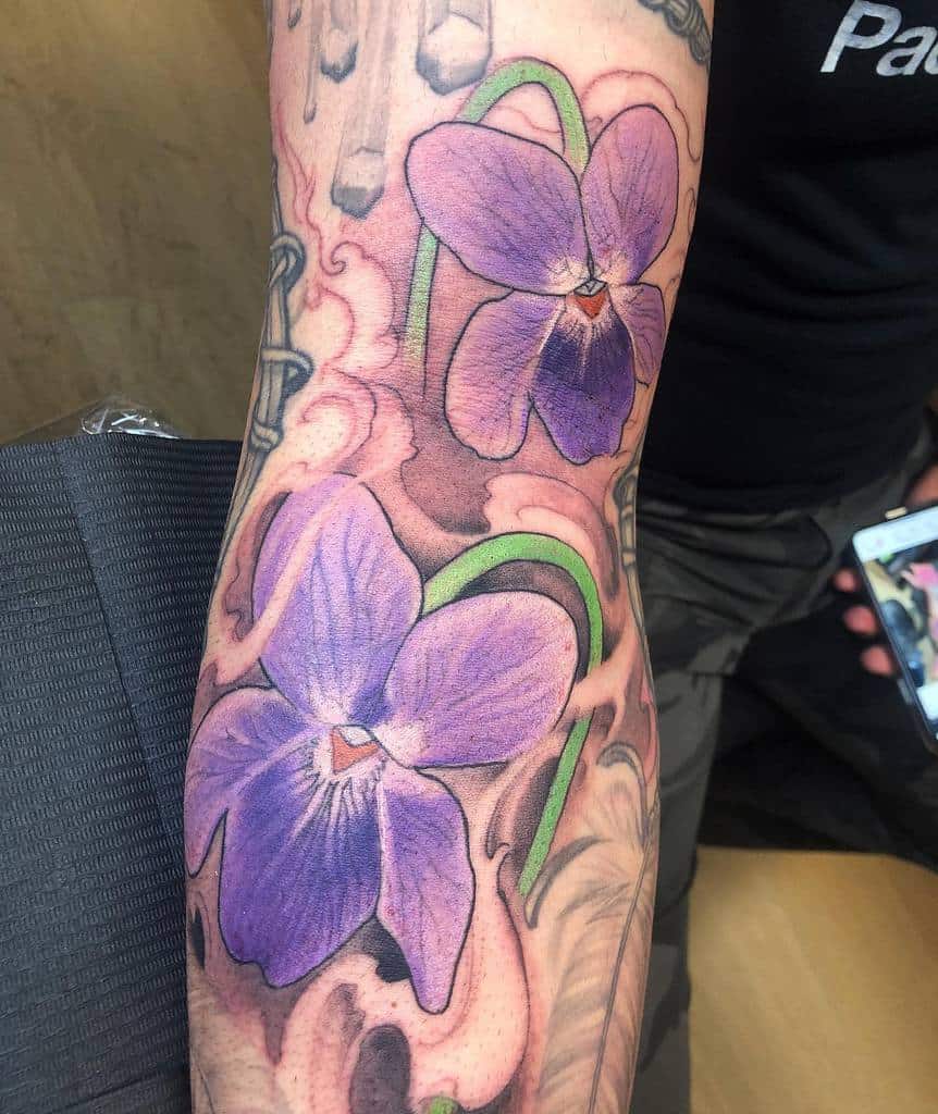 Bold violet flower tattoo with intricate shading, green stems, and a smoky pink background for a striking contrast