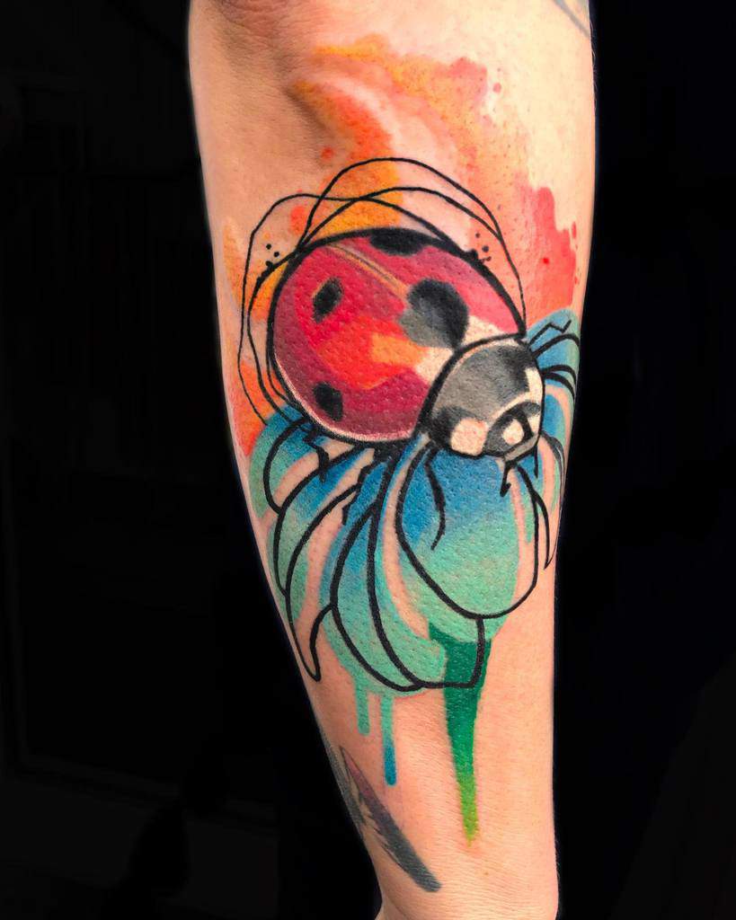 Vibrant watercolor ladybug tattoo with bold red, blue, and orange splashes, featuring a delicate floral design and artistic ink splatters.