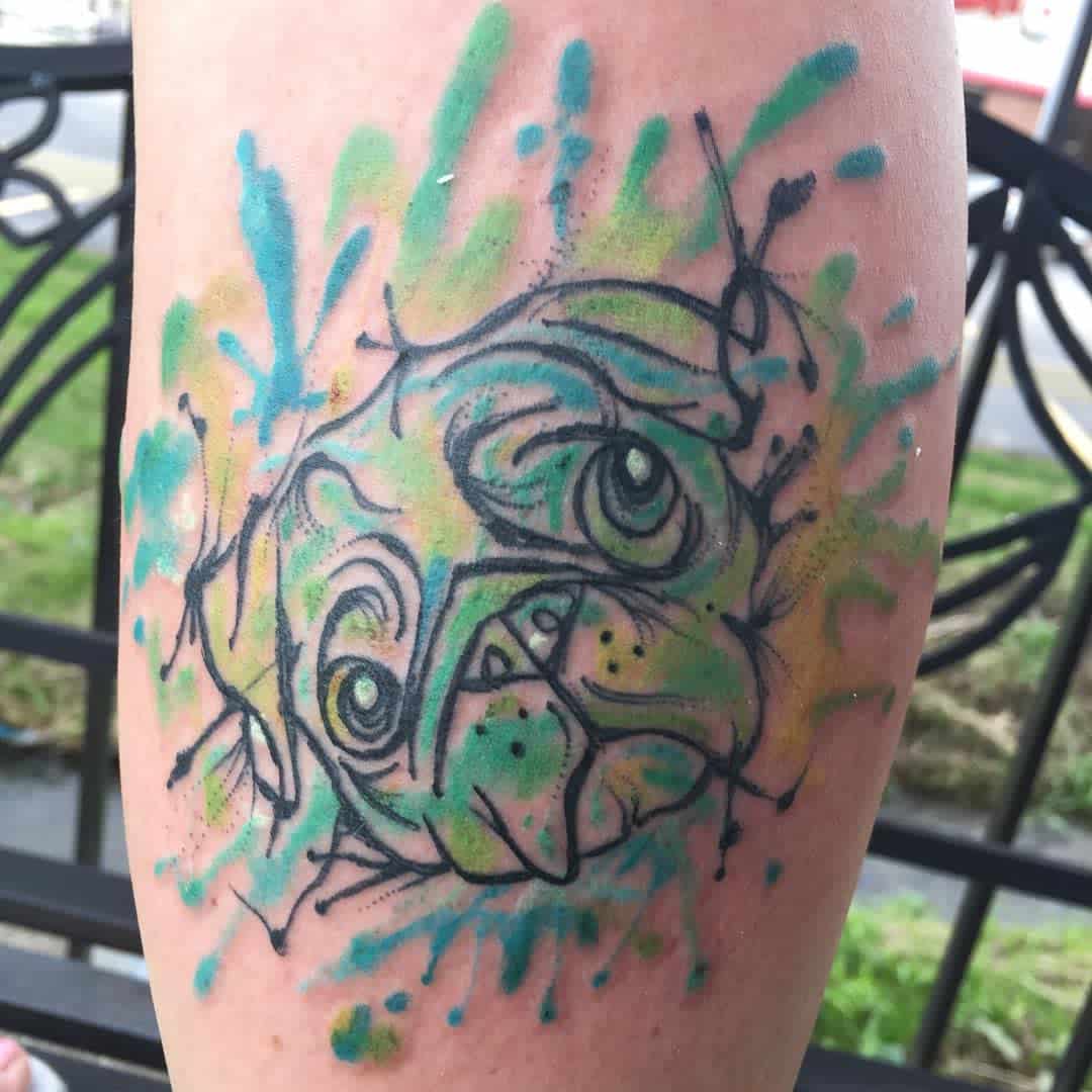 Watercolor-style tattoo of a dog's face with splashes of green, yellow, and blue ink creating an abstract effect on skin