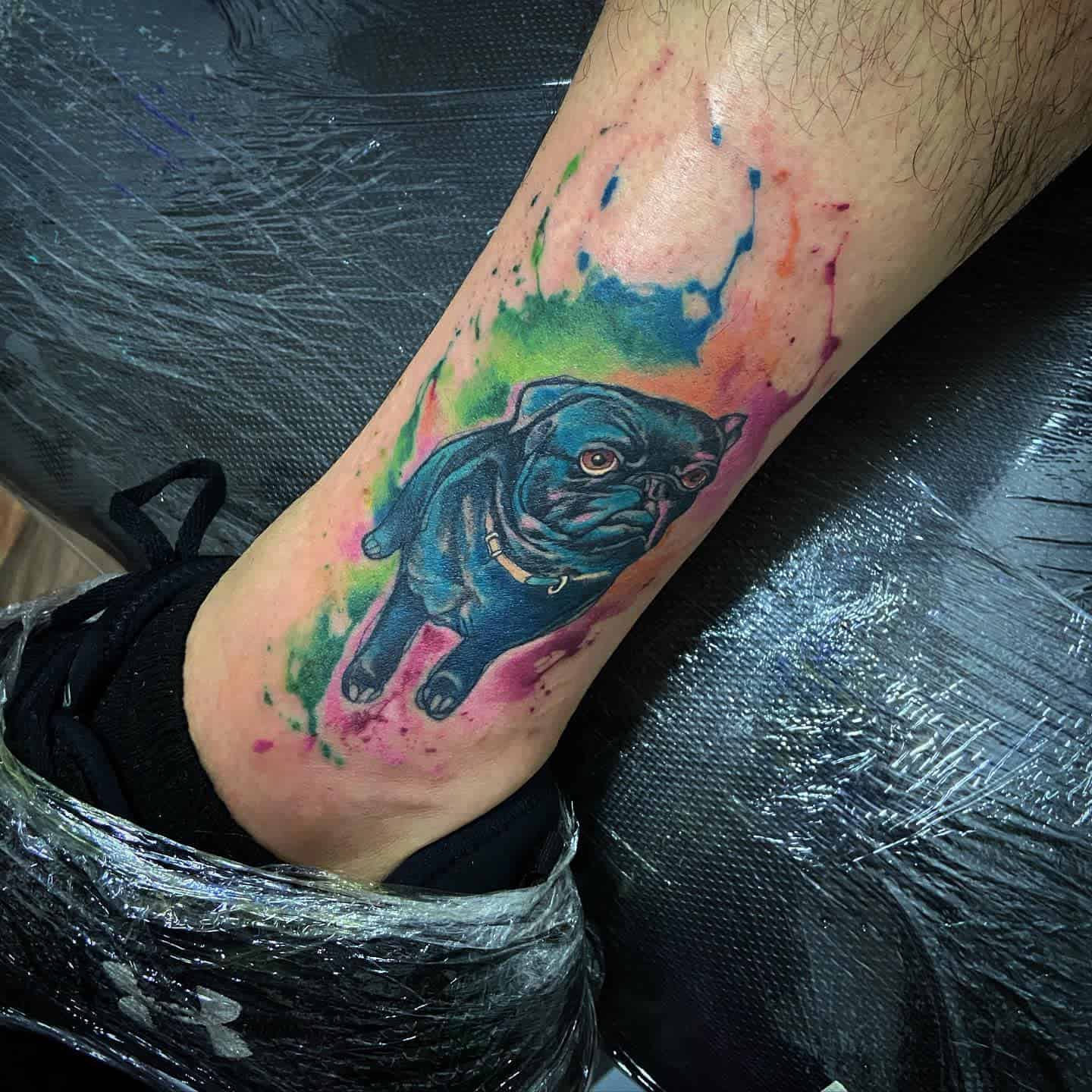 A vibrant tattoo of a pug walking, surrounded by a burst of colorful watercolor paint in shades of blue, green, and pink