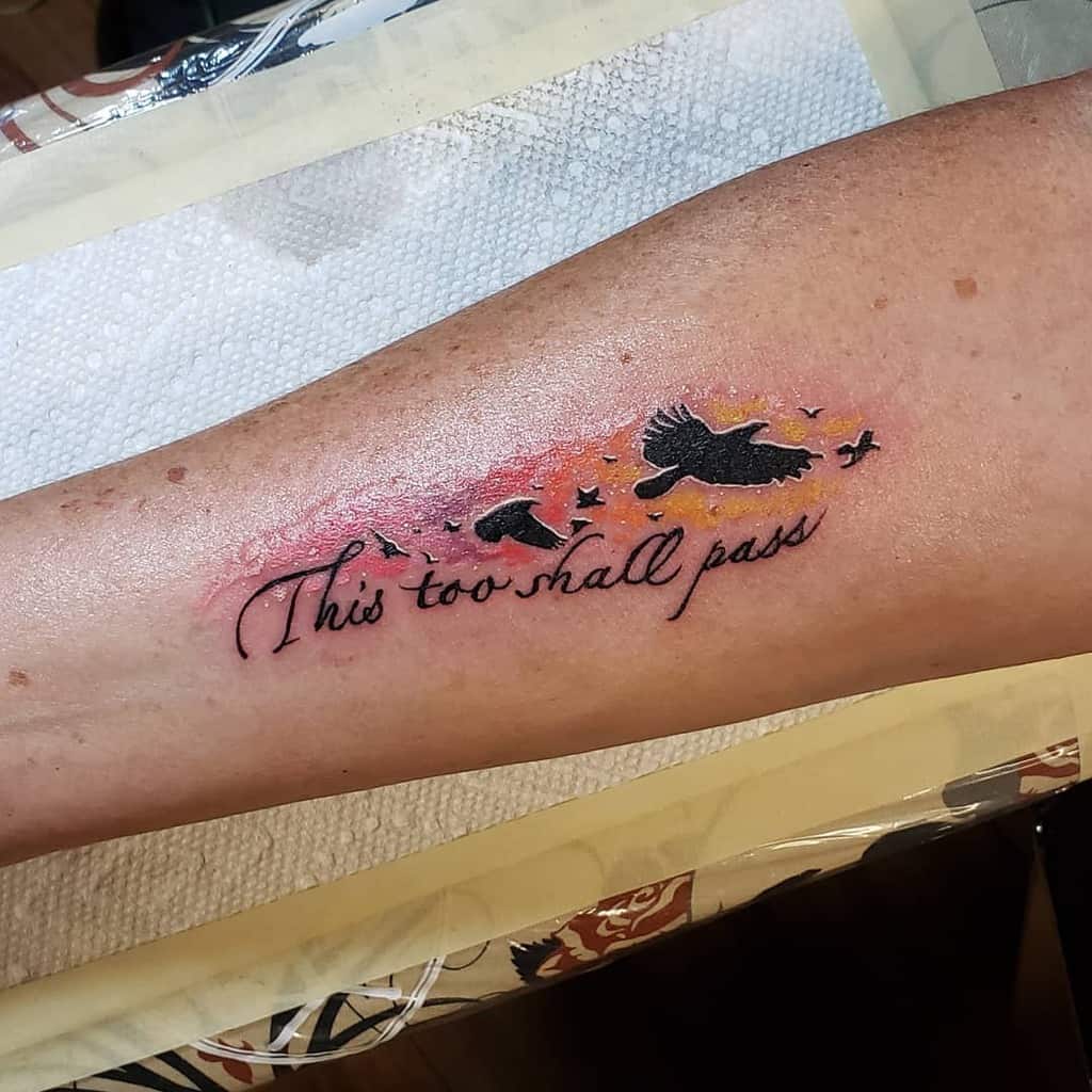 A "This Too Shall Pass" tattoo in cursive with flying birds and a colorful background, on a person's forearm.