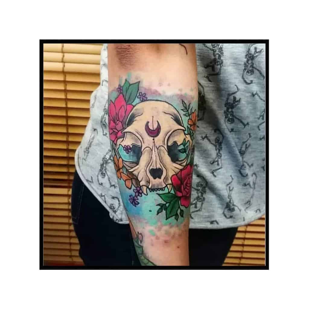 Tattoo of a cat skull adorned with colorful flowers and a crescent moon on an upper arm