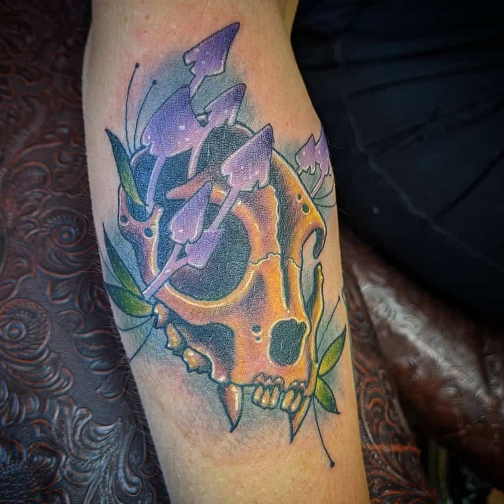 A colorful cat skull tattoo with vibrant mushrooms and leaves adorns the forearm
