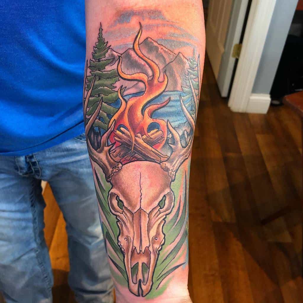 Forearm tattoo featuring a deer skull with antlers, accented by flames, trees, and mountains in the background