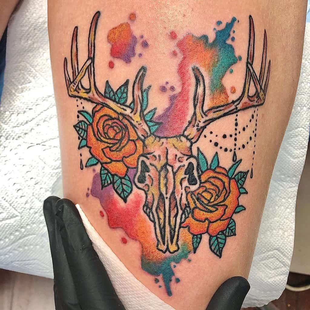 Tattoo of a deer skull with antlers, encircled by vibrant orange roses and striking watercolor splashes