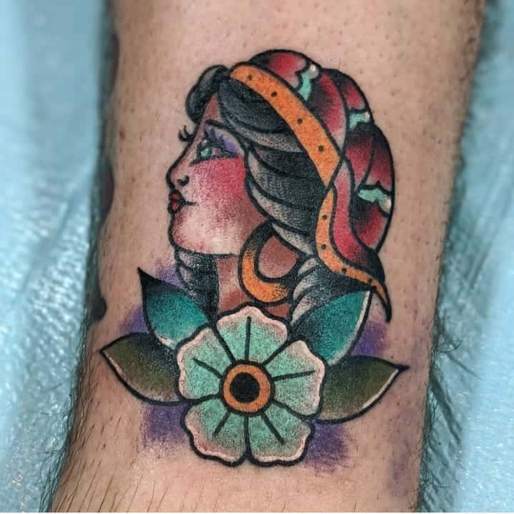 Traditional tattoo of a gypsy woman in profile with a red headscarf, bold shading, and a colorful flower beneath her chin.