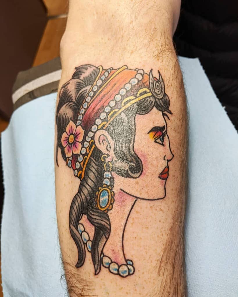 Traditional tattoo of a gypsy woman in profile with an ornate headpiece, pearl jewelry, and floral details on the leg.
