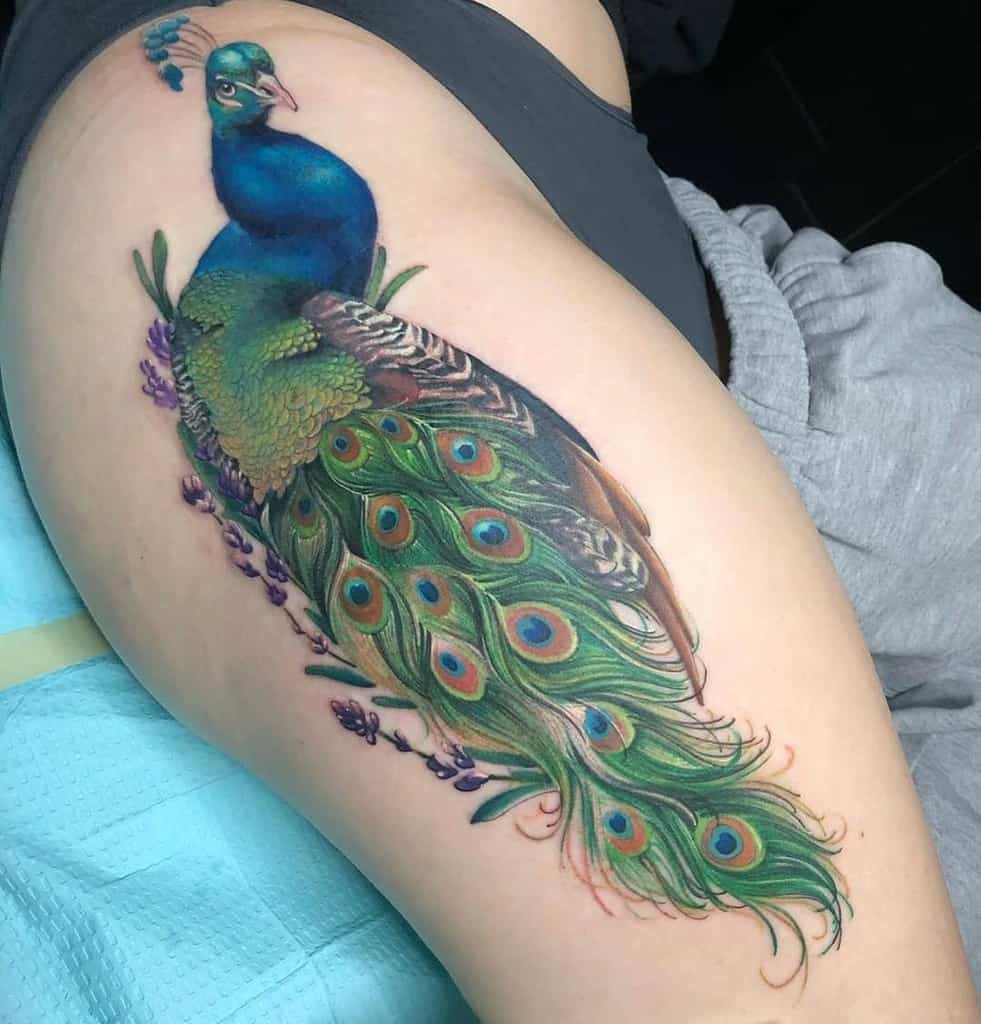 Colorful peacock tattoo on the thigh with vibrant blues and greens in the feathers, accented with delicate lavender flowers around the tail.