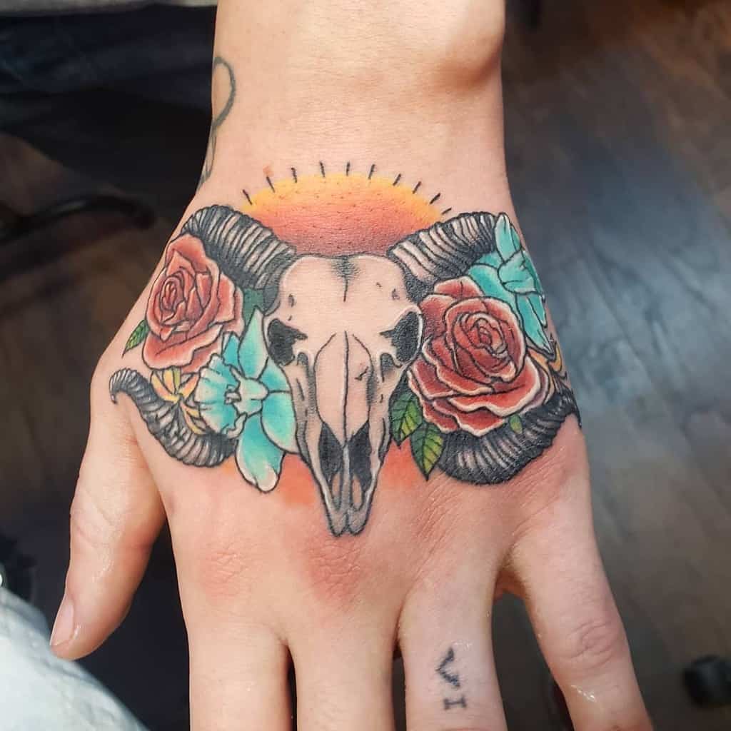 Hand tattoo of a detailed ram skull entwined with roses and turquoise flowers, all framed by a delicate sun