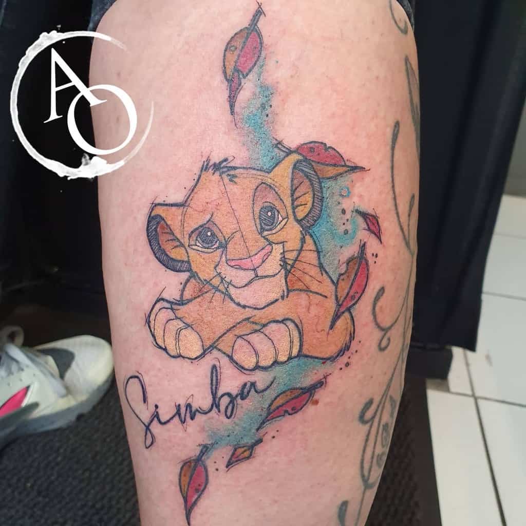 Colorful Simba tattoo featuring a playful cub surrounded by watercolor splashes and autumn leaves, with his name written beneath
