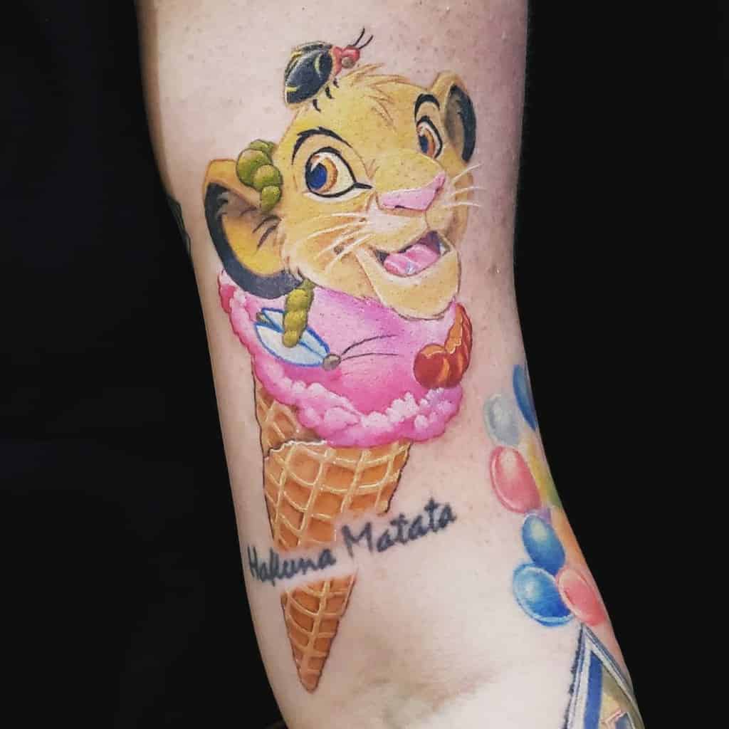 Playful Simba tattoo with a smiling cub on an ice cream cone with strawberry ice cream, accompanied by a ladybug, caterpillar, fly and the phrase 'Hakuna Matata' beneath