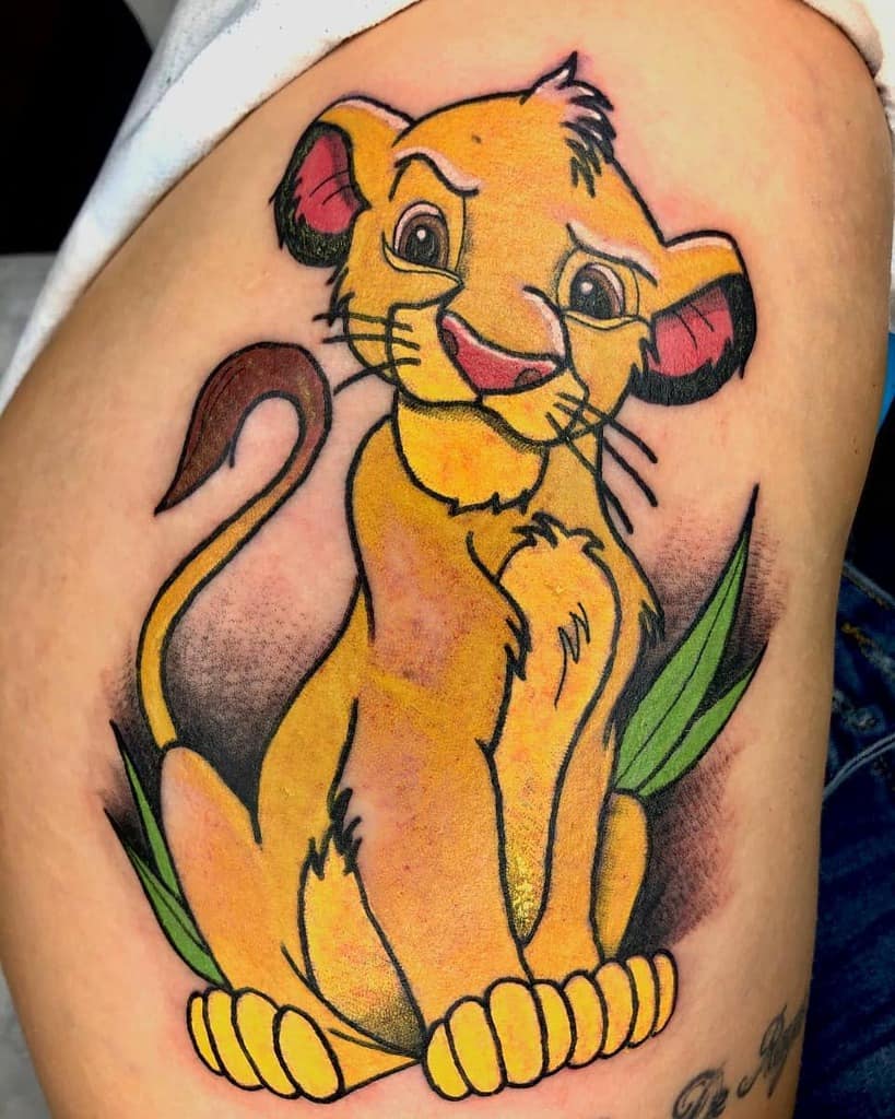 Vibrant and playful tattoo of Simba as a cub sitting with a cheerful expression, surrounded by green leaves for a lively touch