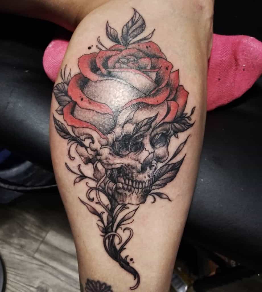 Tattoo of a detailed skull merging into a red rose, with intricate shading and flowing ornamental elements on the leg.