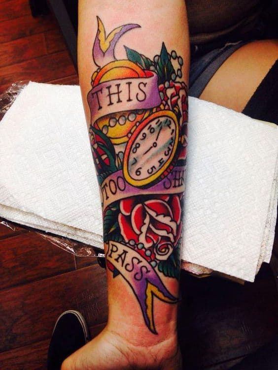 This image displays a vibrant and colorful tattoo on the forearm with the phrase "THIS TOO SHALL PASS" surrounded by roses and a clock. The tattoo features bold lines and bright colors, creating a vivid design.