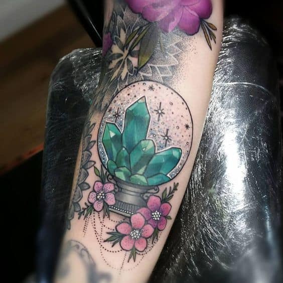 Elegant tattoo of a crystal ball with green gemstones, surrounded by pink flowers and delicate dot work on the forearm.