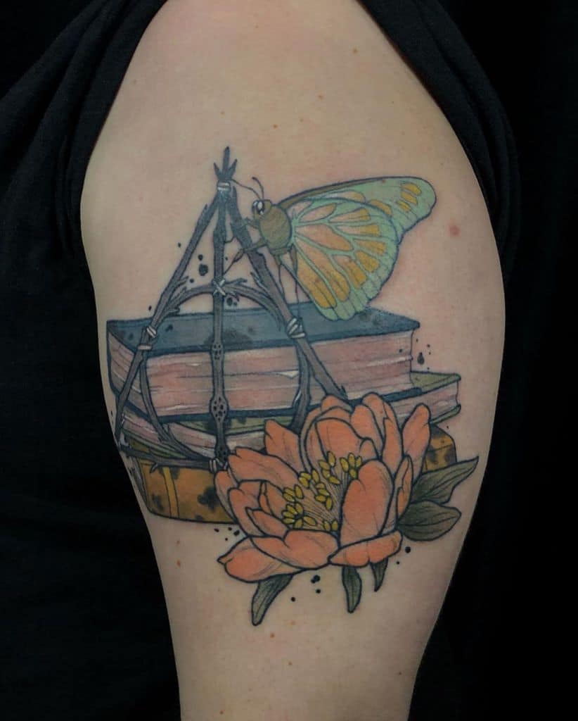 Deathly Hallows tattoo featuring stacked books, a delicate butterfly, and a blooming flower in soft colors for a magical, literary design.