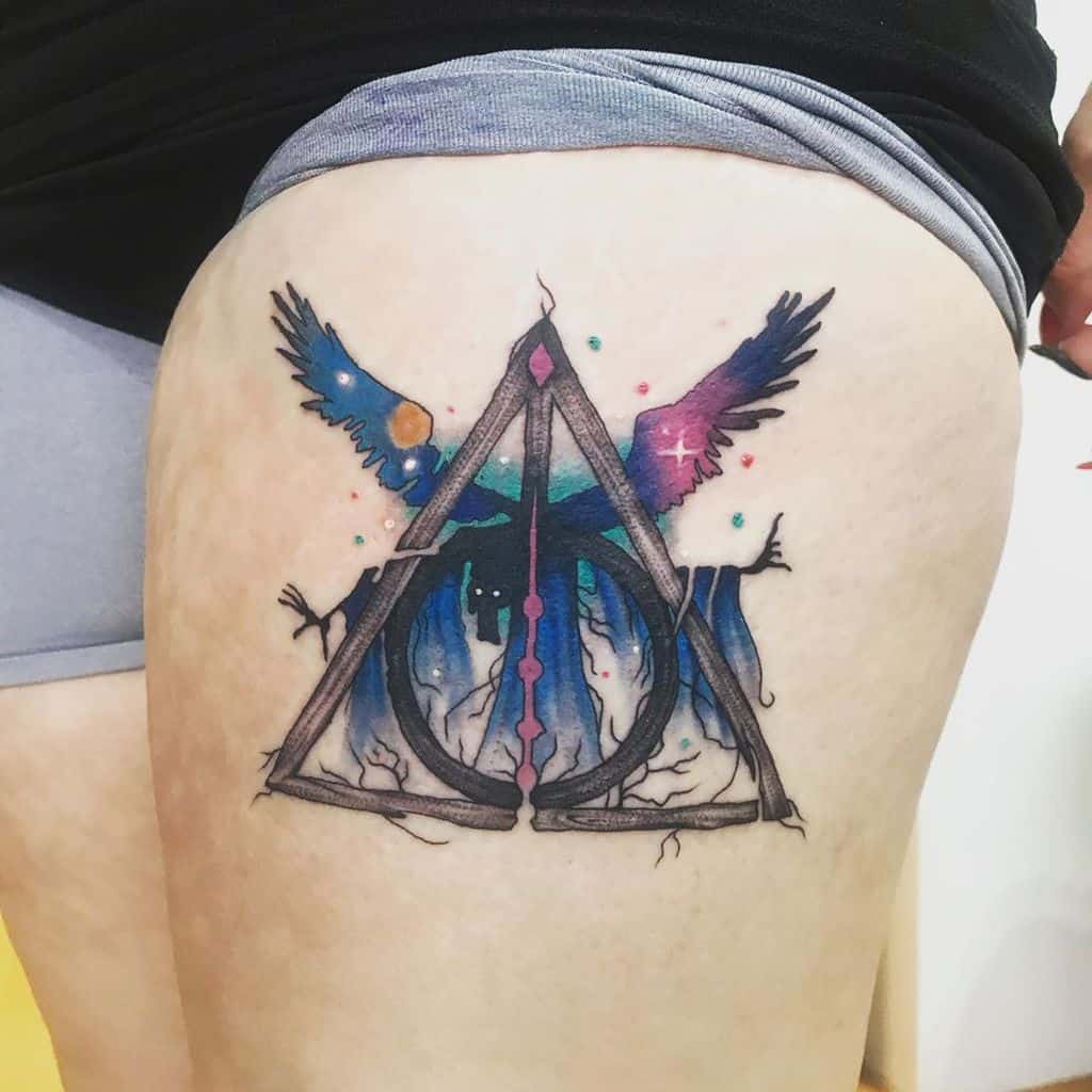 Deathly Hallows tattoo on the thigh with cosmic watercolor effects, a dark forest, and wings blending into a vibrant galaxy design.