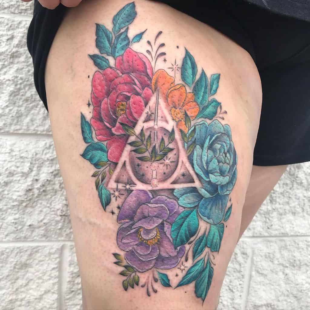 Vibrant Deathly Hallows tattoo on the thigh, surrounded by colorful flowers in shades of red, blue, purple, and orange with magical accents.